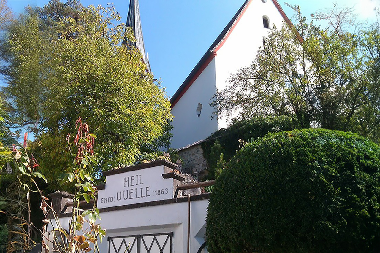 Image of the Mehrn Spring in Tyrol, Austria. Showing image of church and mineral spring right below altar of church. Historic Spring is surrounded by trees and bushes